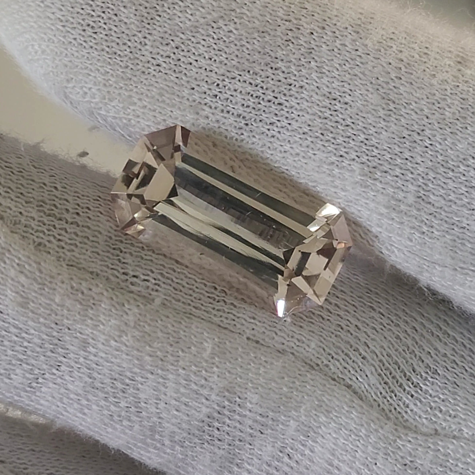 A rectangular emerald-cut clear gemstone set in a silver-toned metal band, shown resting on a white textured fabric surface.