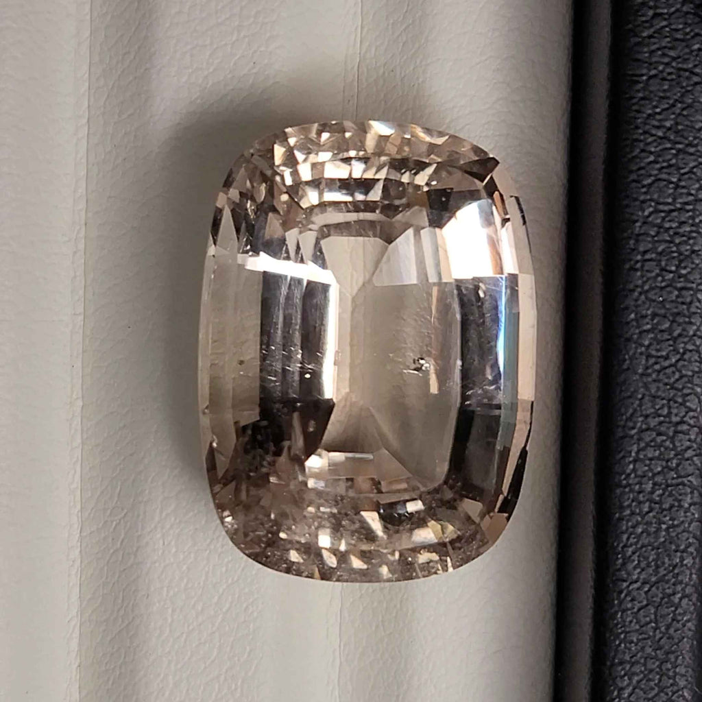 A cushion-cut clear gemstone with a smoky brown tint, photographed against a white paneled background with a black textured strip on the left.