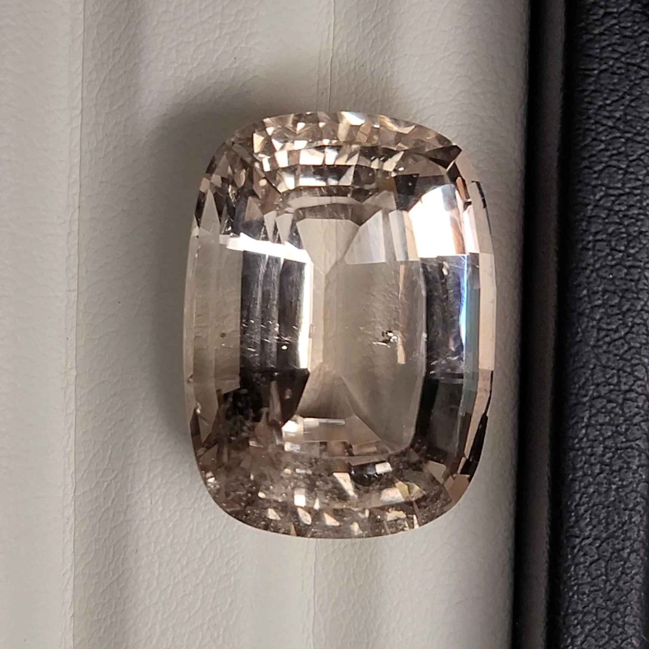 A cushion-cut clear gemstone with a smoky brown tint, photographed against a white paneled background with a black textured strip on the left.