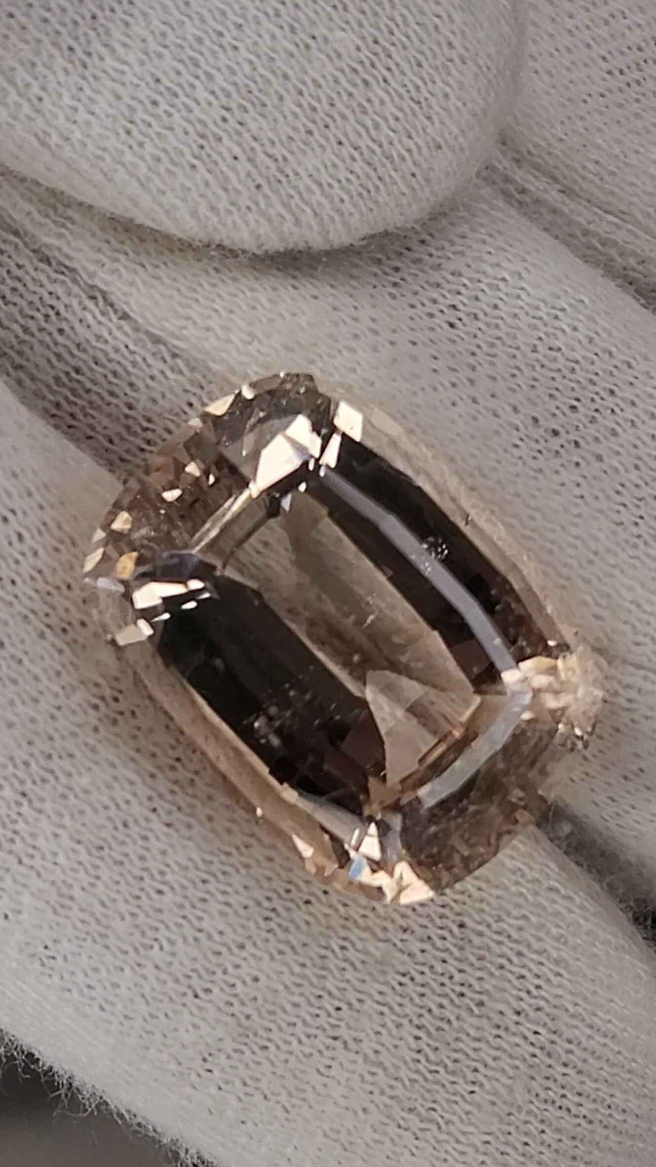 A rectangular cushion-cut clear gemstone with a pinkish-brown tint, held between white fabric gloves.